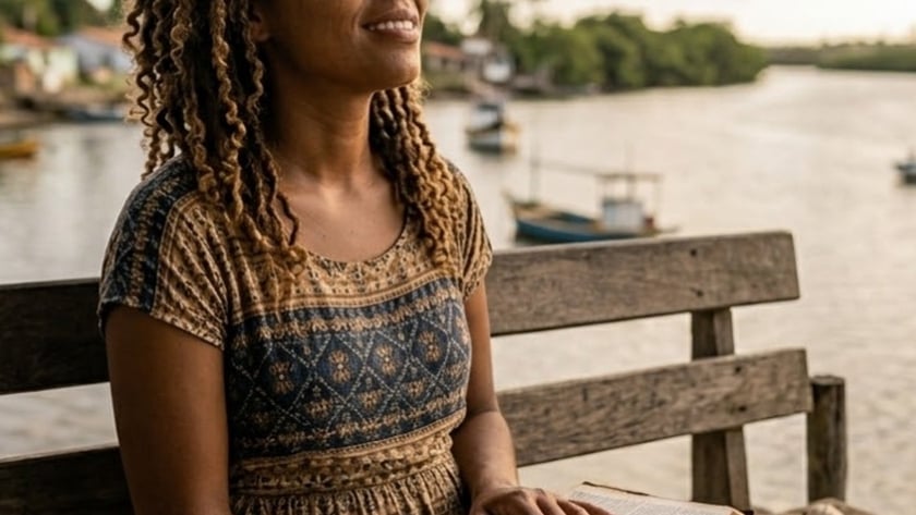 Mulher de cabelo cacheado sentada num banco de madeira com um livro aberto, diante de um rio.