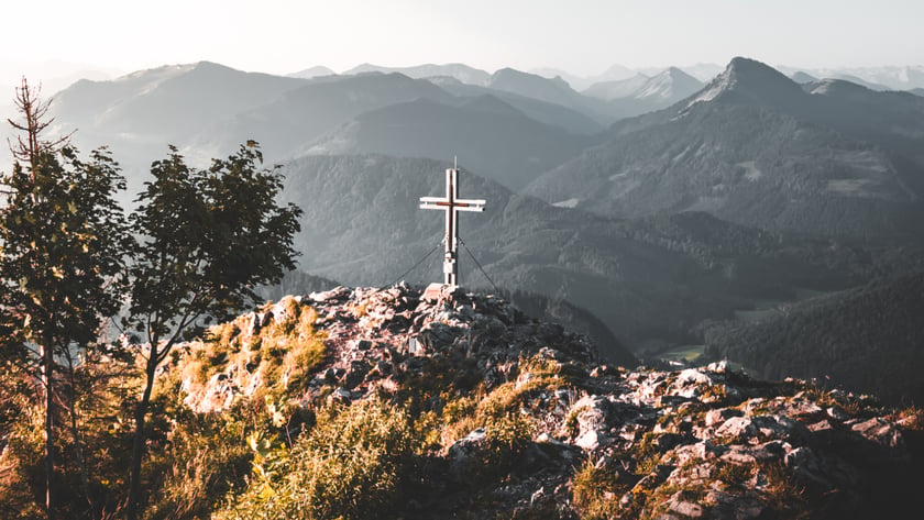 Cruz de metal no topo rochoso de uma montanha, com cadeias de montanhas ao fundo.