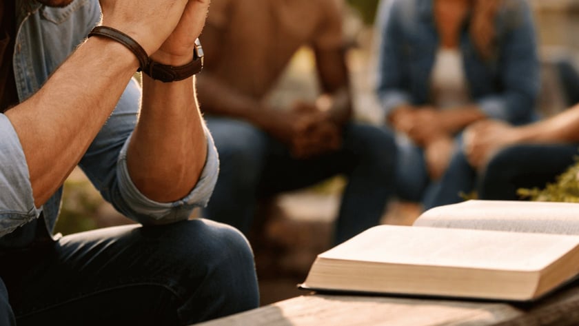 Homem de cabeça baixa com as mãos unidas, perto de uma Bíblia aberta; ao fundo