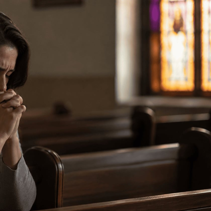 Mulher de mãos unidas junto ao rosto, sentada em um banco de igreja.