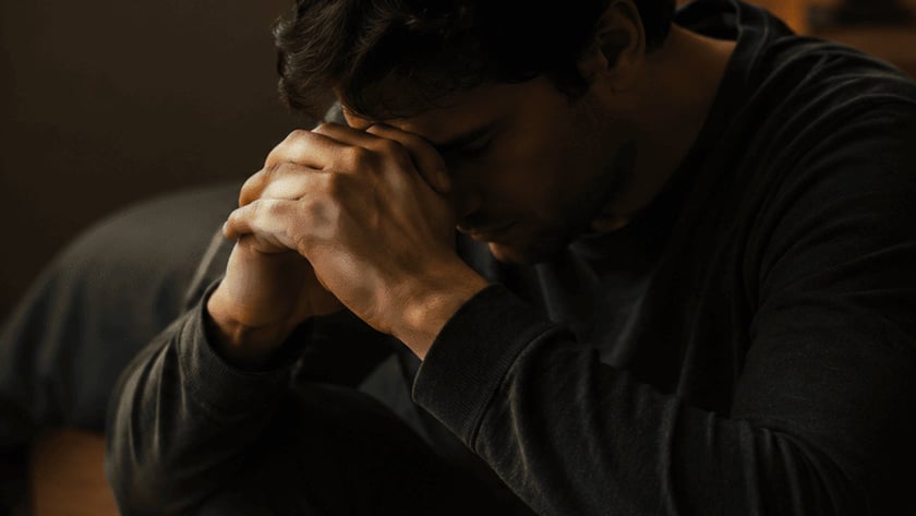 Homem de cabelo escuro e blusa preta sentado num quarto, com a cabeça apoiada nas mãos unidas.
