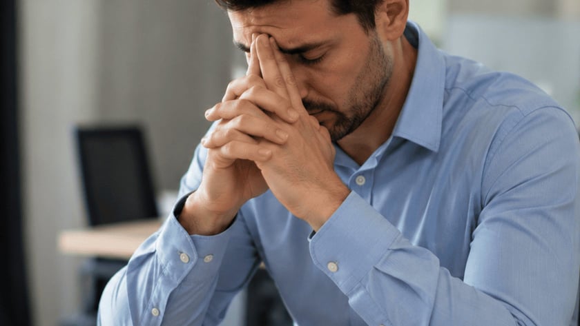 Homem de camisa azul apoia o rosto nas mãos diante de um notebook, em escritório.