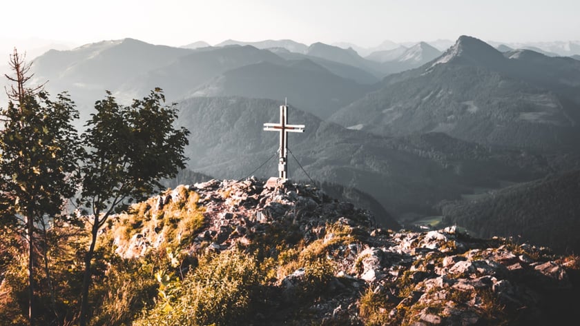Cruz de metal no topo rochoso de uma montanha, com cadeias de montanhas ao fundo.
