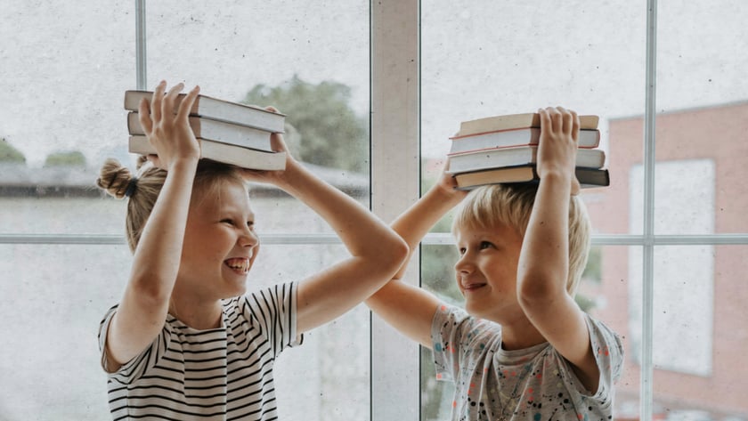 Duas crianças sentadas no parapeito de uma janela, cada uma com uma pilha de livros na cabeça.