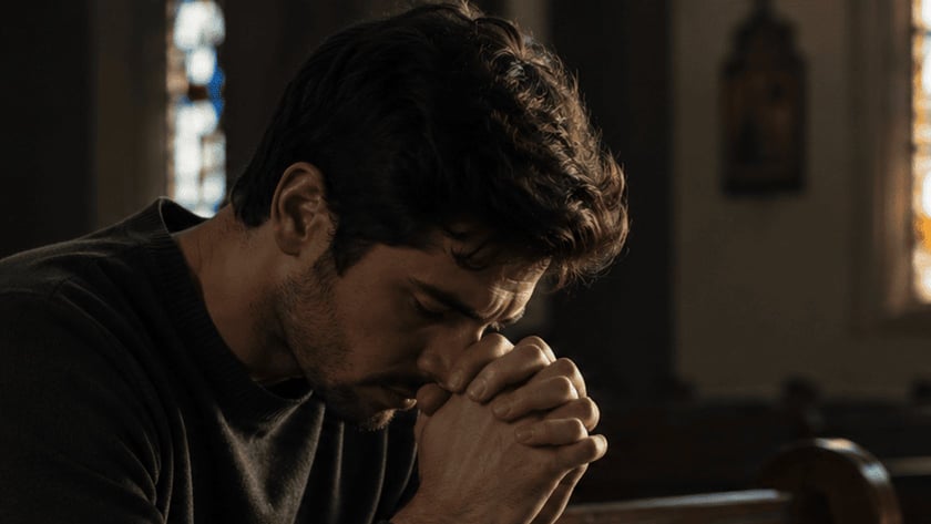 Homem de cabelo escuro, com as mãos unidas junto ao rosto, sentado em um banco de igreja.