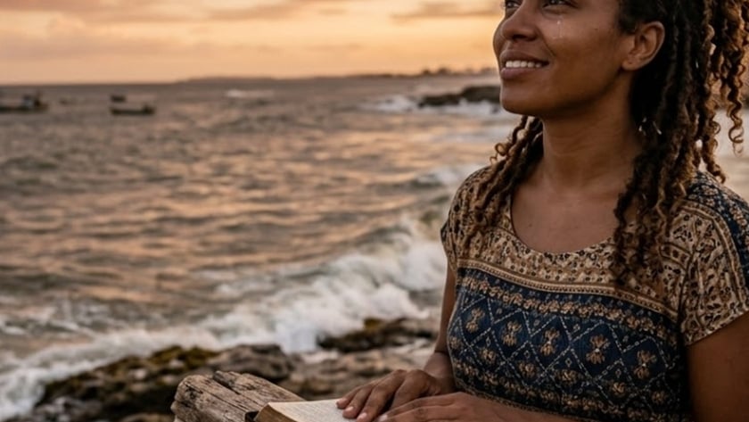 Mulher de cabelo cacheado segura uma Bíblia aberta sobre um tronco, à beira-mar, ao entardecer.