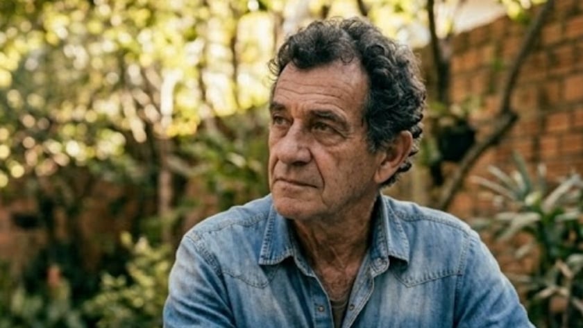 Homem de camisa jeans sentado a uma mesa em um quintal com plantas.