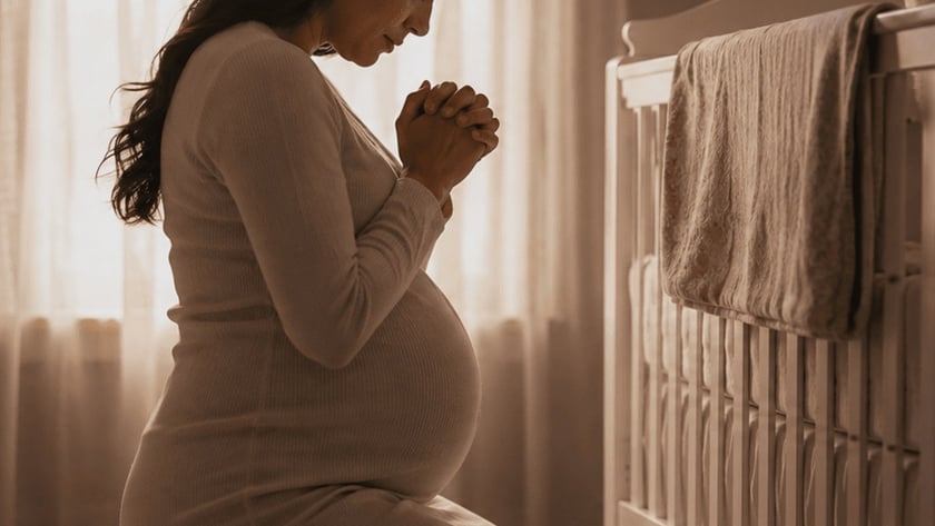 Mulher grávida ajoelhada com as mãos unidas, de olhos fechados, em um quarto de bebê.