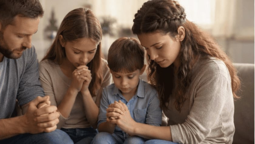Mulher, menina e menino sentados juntos, de olhos fechados e mãos unidas, rezando.