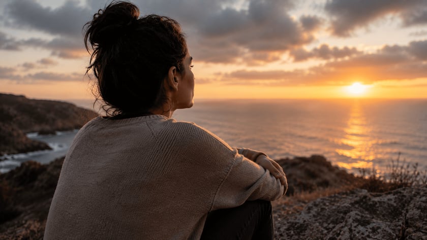 Mulher de coque e suéter bege, vista de costas, observa o mar do alto de um penhasco.
