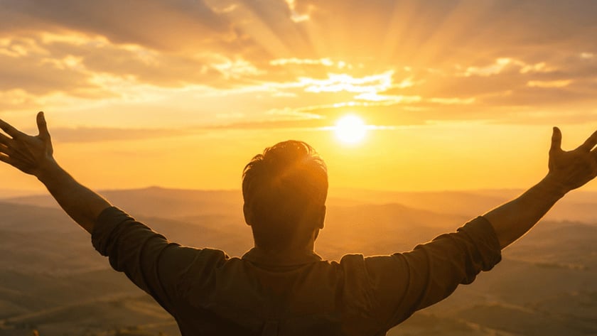 Homem de costas, com os braços abertos, olha o pôr do sol sobre colinas.