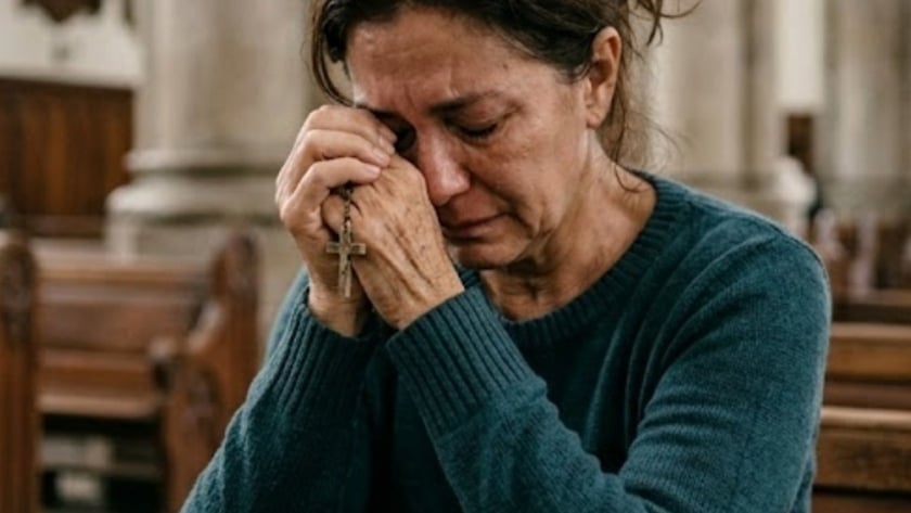 Mulher de suéter azul segura um terço junto ao rosto, sentada num banco de igreja.