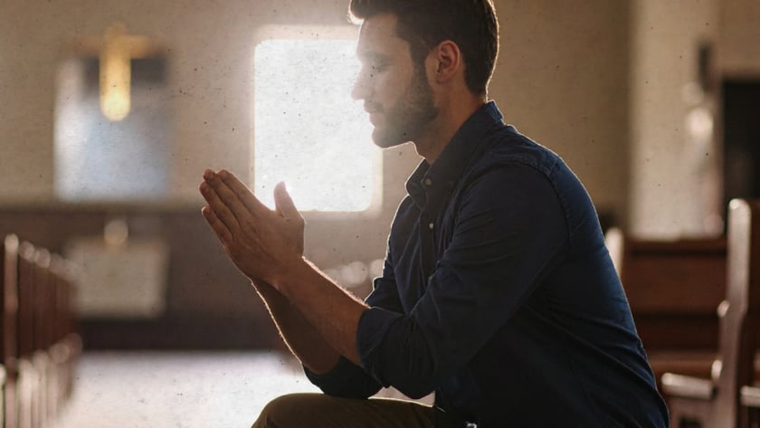 Homem ajoelhado de mãos juntas, rezando entre os bancos de uma igreja.