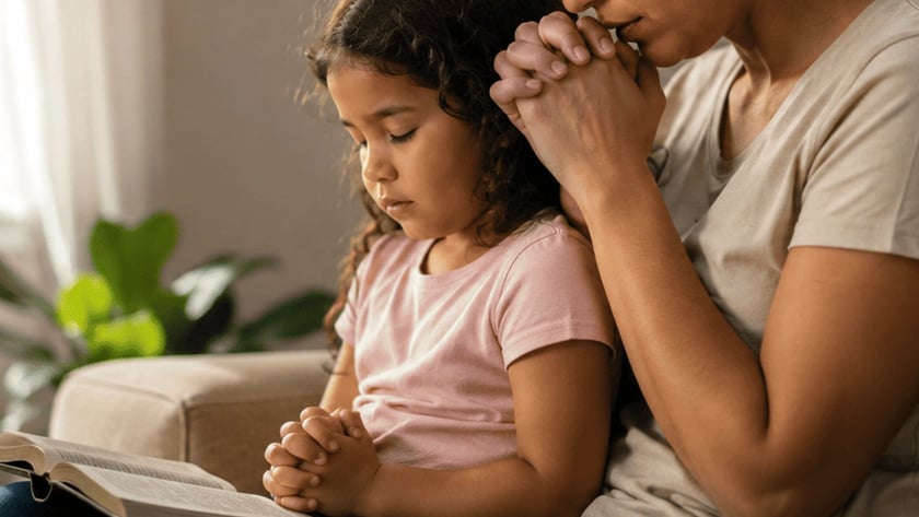Mulher e menina de olhos fechados e mãos juntas, rezando no sofá com uma Bíblia aberta.