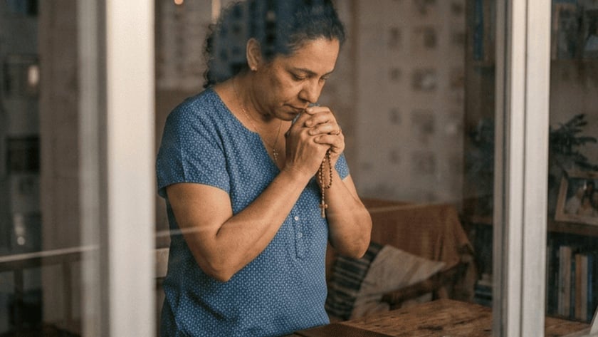 Mulher de blusa azul reza com um terço junto à janela, diante de uma Bíblia e uma vela.