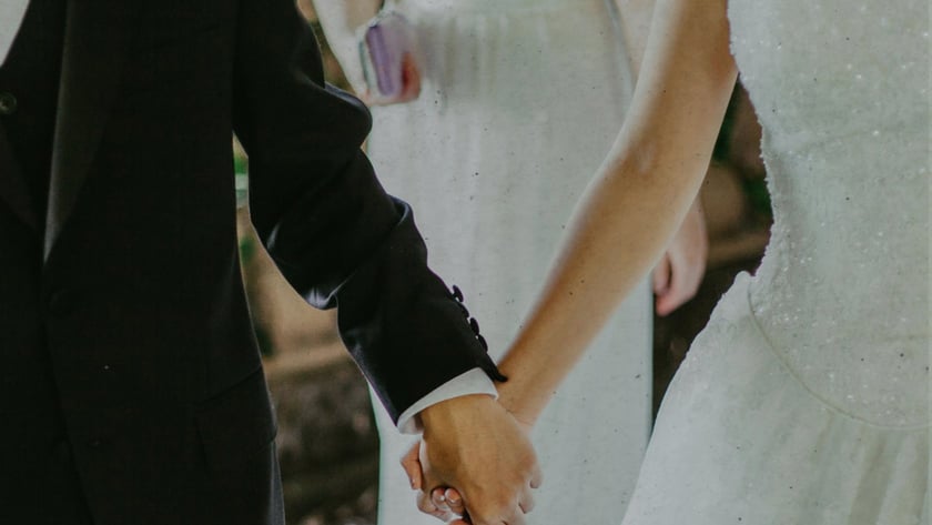 Noiva de vestido branco e noivo de terno escuro andam de mãos dadas.