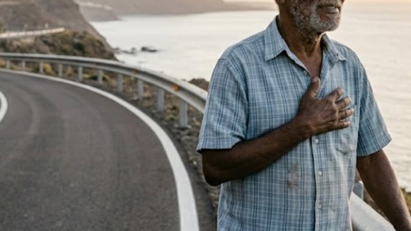 Homem idoso de camisa xadrez, com a mão no peito, ao lado de uma estrada à beira-mar.