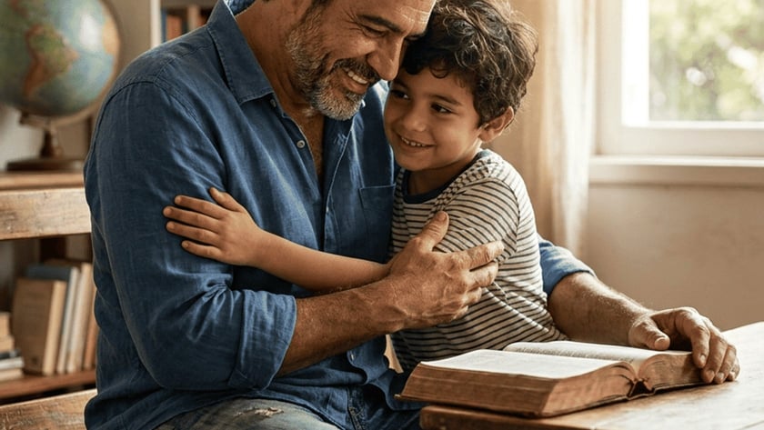 Homem sorridente abraça um menino ao lado de uma mesa com livro aberto.