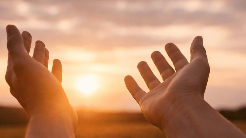 Duas mãos abertas erguidas em direção ao sol poente sobre um campo.