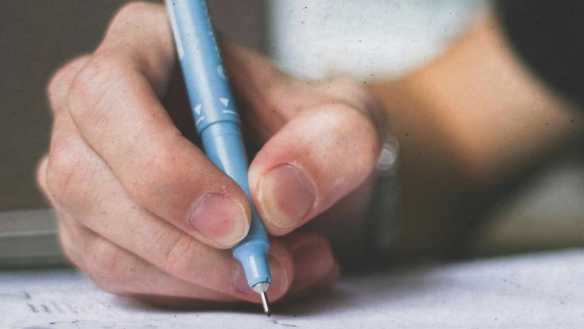 Mão segurando uma caneta azul-clara e escrevendo em um caderno aberto sobre a mesa.