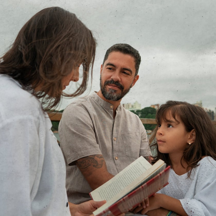 Homem de barba, mulher e menina conversam ao ar livre segurando um livro aberto.