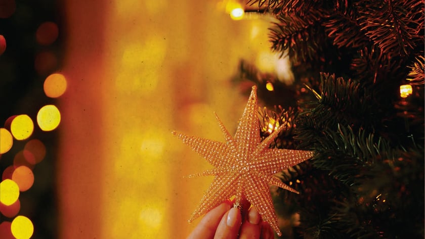 Duas mãos seguram uma estrela dourada de contas junto a uma árvore de Natal.