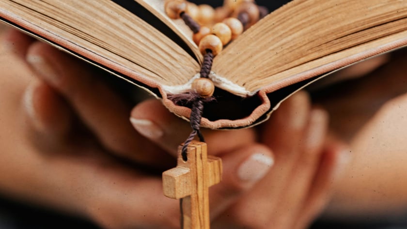 Mãos seguram um livro aberto com um terço de contas de madeira e cruz.