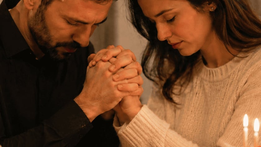 Casal de mãos dadas e cabeça baixa à mesa, com bolo de velas acesas.