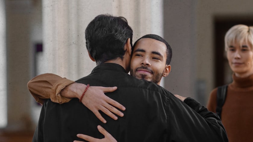 Dois homens se abraçam em uma igreja, enquanto um jovem loiro os observa ao fundo.
