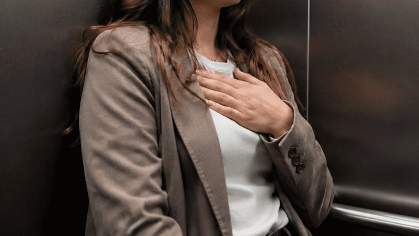 Mulher de blazer marrom encostada na parede de um elevador, com a mão no peito.