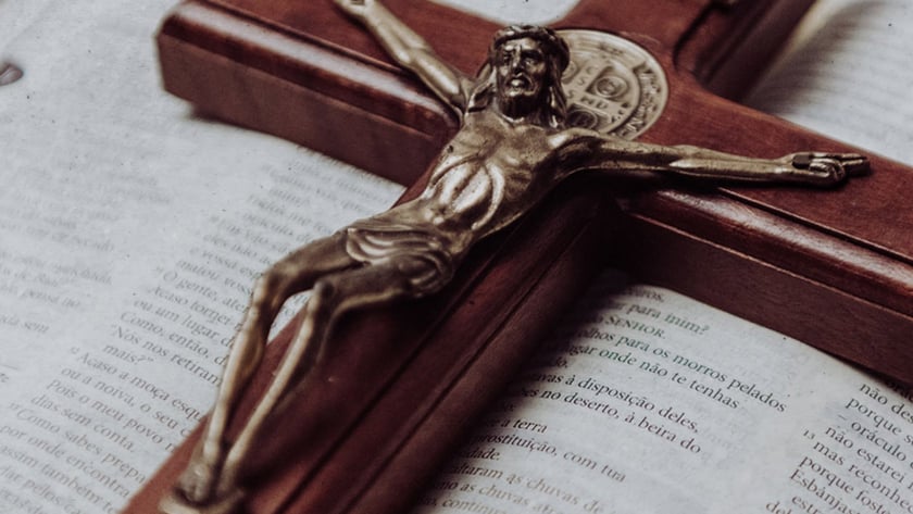 Crucifixo de madeira com Cristo metálico sobre Bíblia aberta, com tecido roxo ao fundo. "INRI"