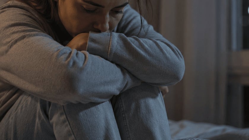 Mulher sentada na cama abraça os joelhos e apoia o rosto nos braços.
