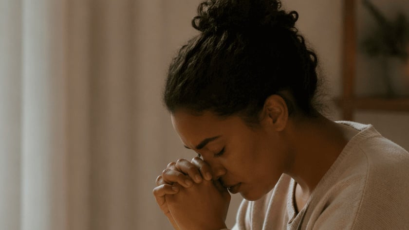 Mulher de olhos fechados e mãos postas sobre uma Bíblia aberta, à mesa, ao lado de vela e caneca.