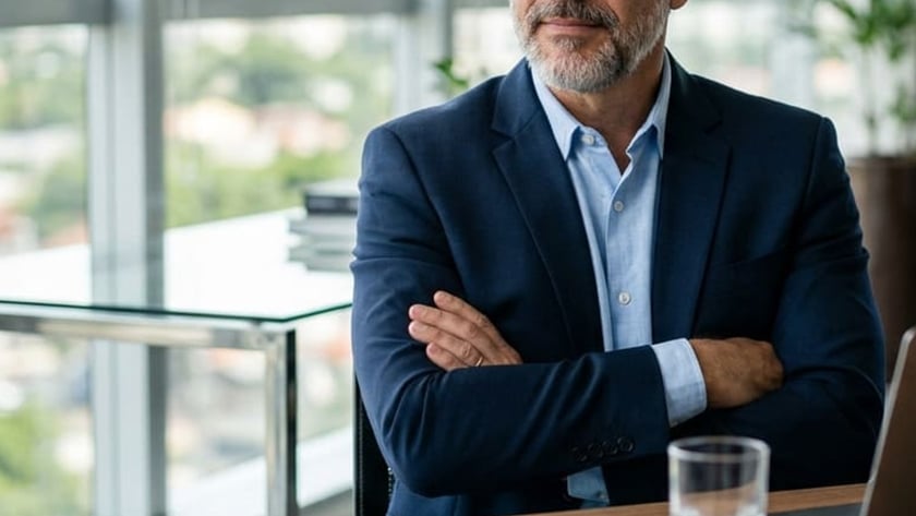 Homem grisalho de blazer azul e braços cruzados ouve outra pessoa em escritório envidraçado.