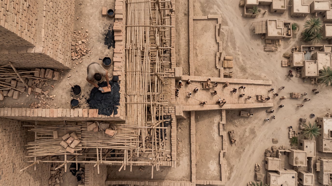 Operários constroem uma muralha de tijolos com andaimes de bambu, perto de casas de barro.