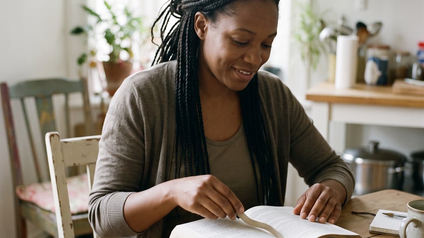 Mulher de tranças lendo um livro aberto sobre uma mesa de madeira, em uma cozinha.