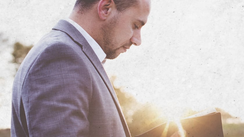 Homem de blazer cinza olha para um livro aberto em suas mãos, ao ar livre.