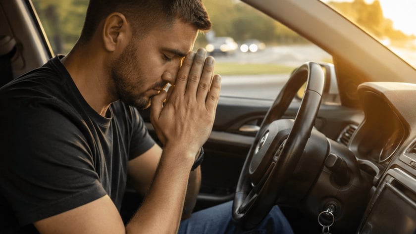 Homem de camiseta escura sentado ao volante, com as mãos unidas diante do rosto e olhos fechados.