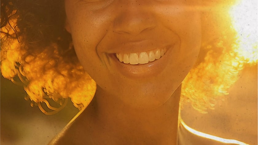 Mulher de cabelo cacheado sorri para a câmera, com o sol atrás dela.
