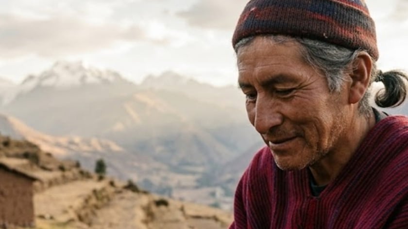 Homem de gorro e poncho vermelho segura sementes nas mãos, em terreno de montanha.