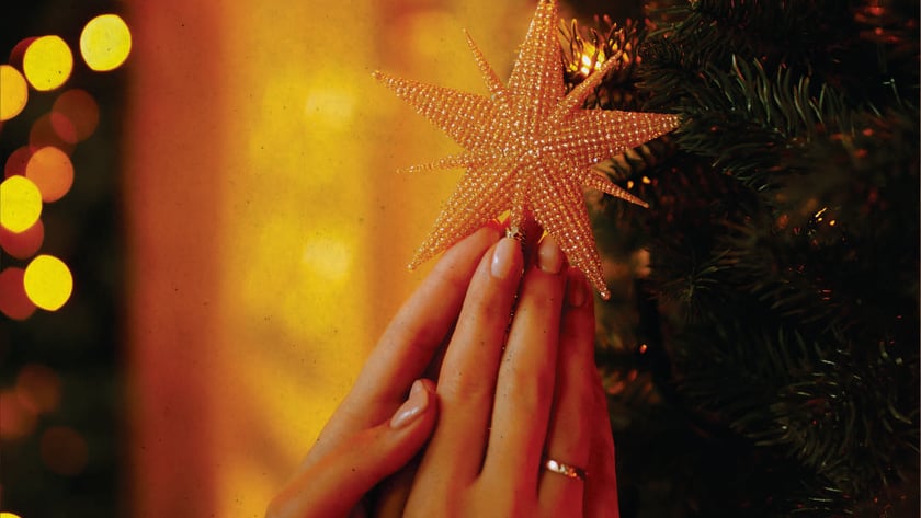 Duas mãos seguram uma estrela dourada de contas junto a uma árvore de Natal.