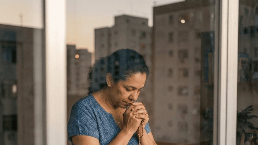 Mulher de blusa azul reza com um terço junto à janela, diante de uma Bíblia e uma vela.