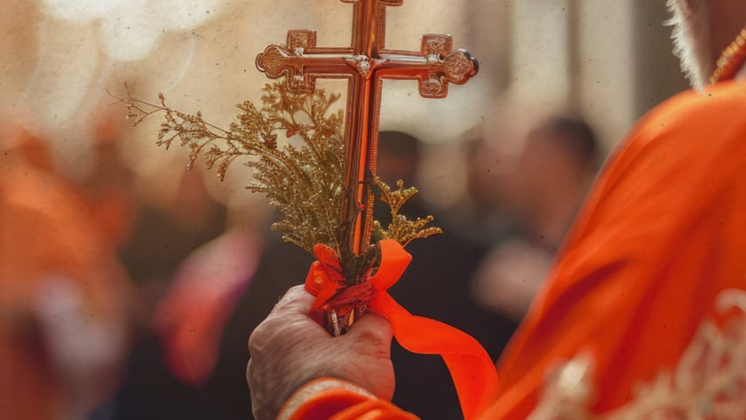 Religioso de vestes laranja segura uma cruz dourada com ramos e fita, diante de uma multidão.