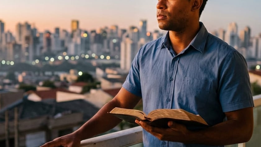 Homem de camisa azul segura um livro aberto em uma varanda, diante de uma cidade.