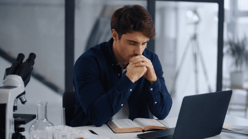 Homem de camisa azul com as mãos unidas junto ao rosto, diante de uma Bíblia aberta e um notebook.