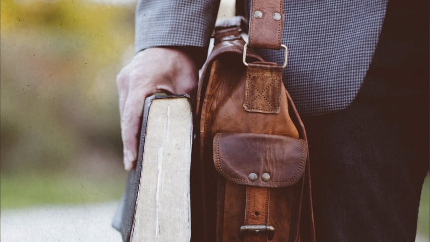 Homem de paletó segura um livro grosso e carrega uma bolsa de couro marrom.