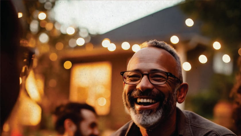 Homem de óculos e barba grisalha rindo em um jardim com luzes ao fundo.