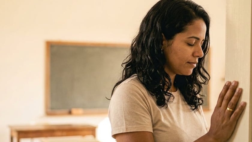 Mulher de cabelo escuro e olhos fechados apoia a mão na parede de uma sala de aula vazia.