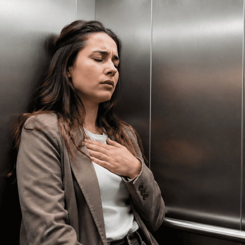 Mulher de blazer marrom encostada na parede de um elevador, com a mão no peito.