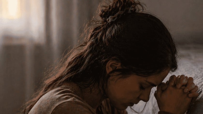 Mulher de cabelo preso ajoelhada ao lado de uma cama, com as mãos unidas junto à testa.